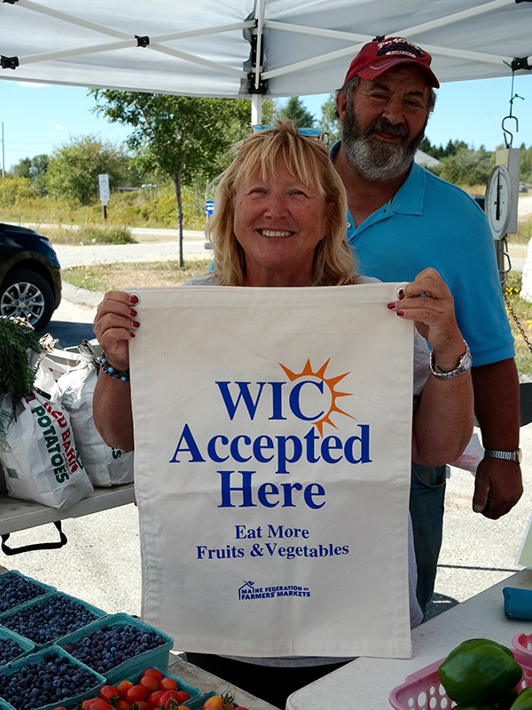 A vendor at a farmers' market smiles as she holds a sign that says "WIC accepted here"